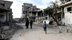 Reuters People, including a child on a crutch, missing the lower half of his left leg, walk past the grey, dusty rubble of houses and buildings destroyed during the war in Al-Bureij in central Gaza