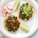 Two california street tacos with lime and radish on a paper plate, surrounded by Mexican drinks and salsa.