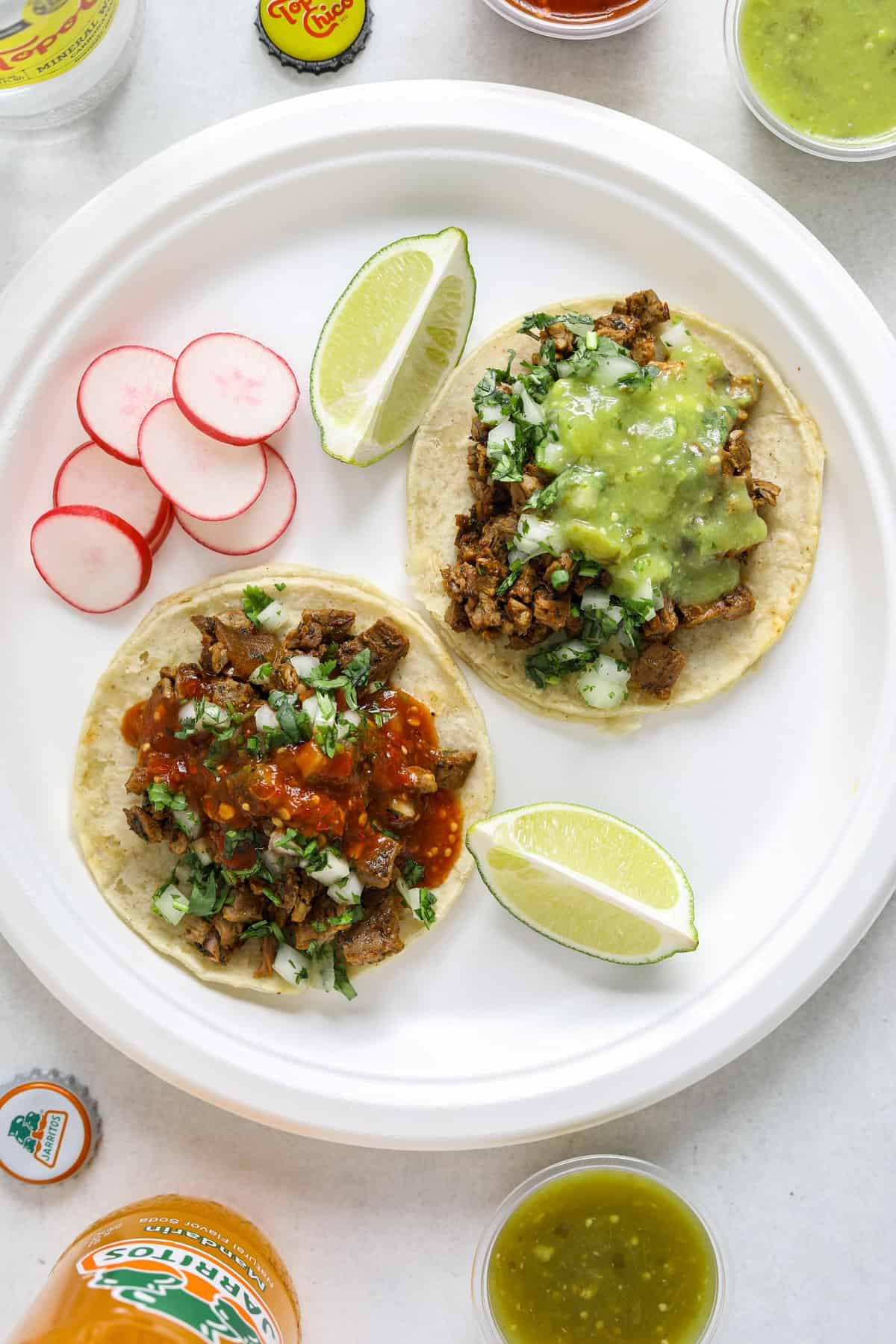 Two california street tacos with lime and radish on a paper plate, surrounded by Mexican drinks and salsa.