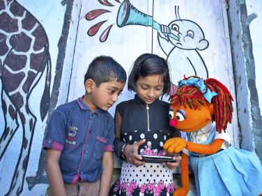 Chamki playing on a tablet with children, encouraging users to contact Sesame Workshop for crisis support.