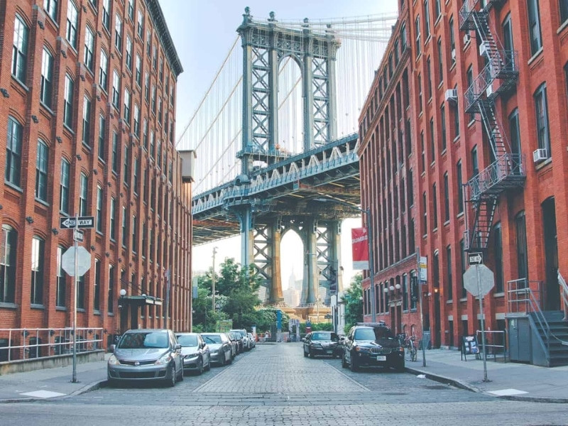 Manhattan Bridge view from Washington Street, framed by red brick buildings in Dumbo, Brooklyn, with cobblestone streets in the foreground