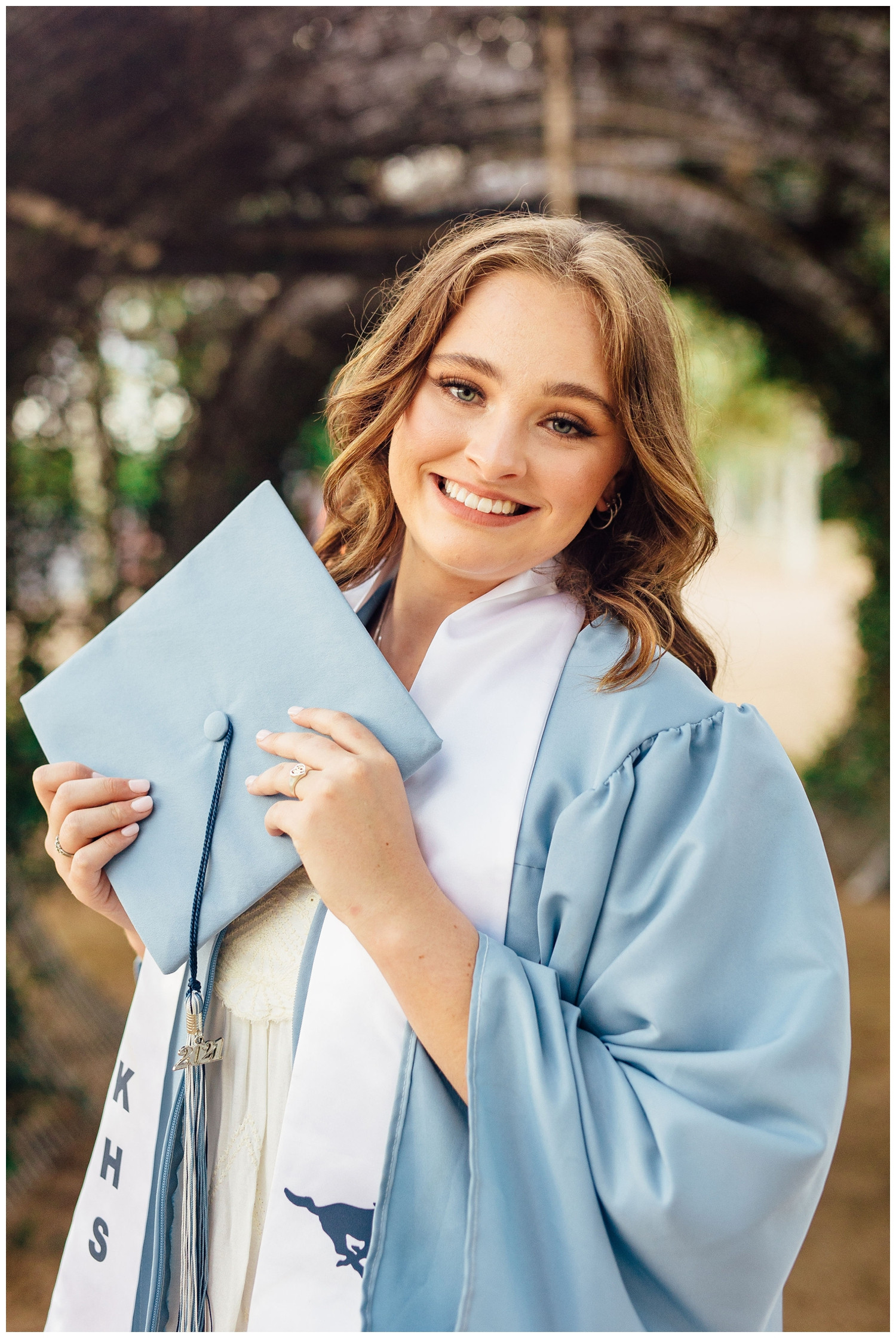 Smiling high school senior in graduation gown holding blue cap, Sabine Street Bridge Houston backdrop