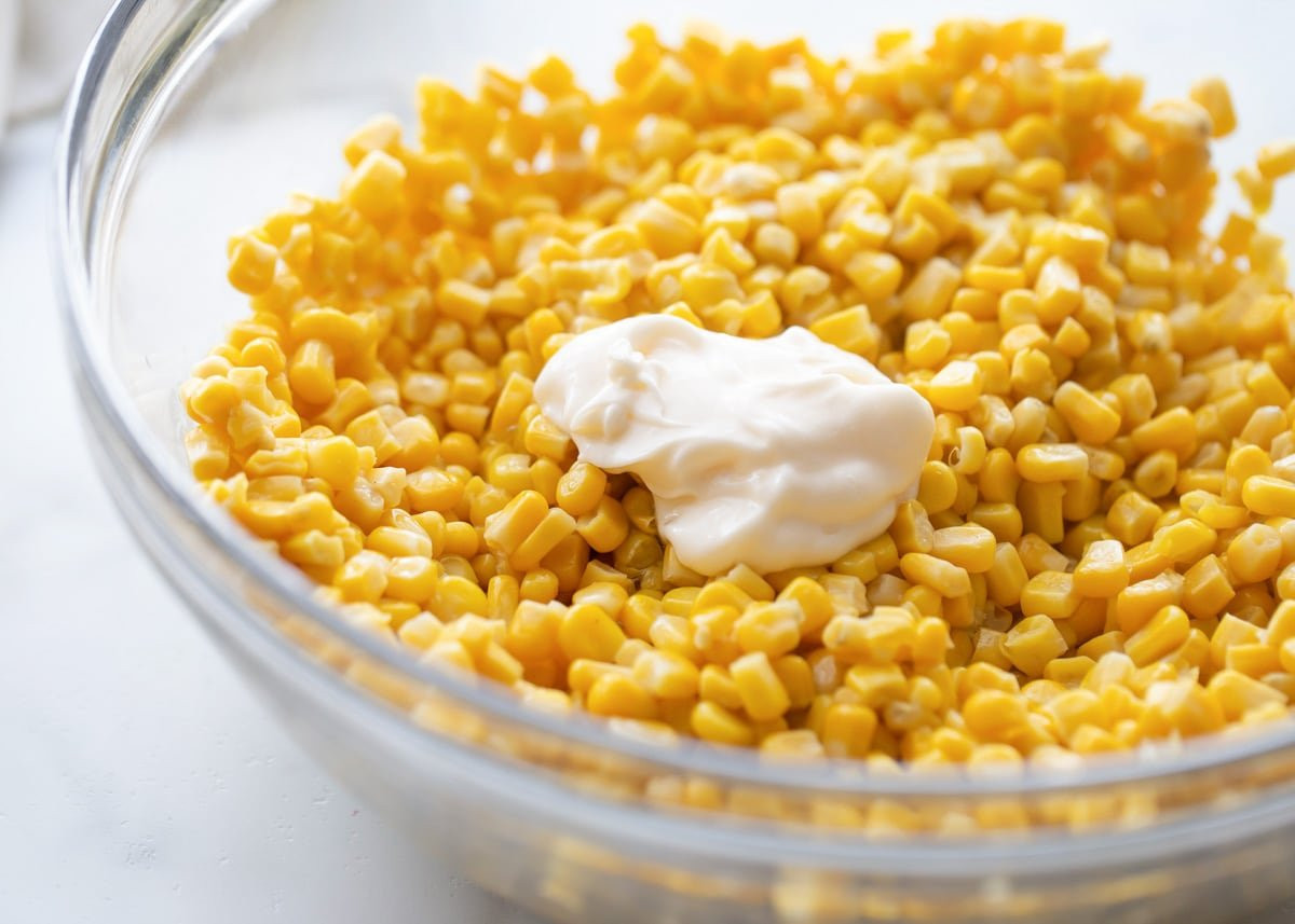 Image displaying corn salad in a glass bowl with mayonnaise being drizzled on top, illustrating the addition of creamy dressing to the corn.