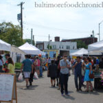 A wide shot of the 32nd Street Farmers Market, showcasing vendor tents and shoppers browsing fresh produce and baked goods under sunny skies, capturing the lively atmosphere of the Waverly Market.