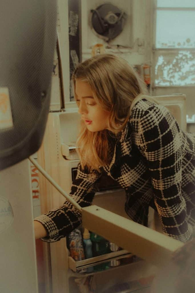A woman with a thoughtful expression, looking into her refrigerator, surrounded by various food items, contemplating what to eat for dinner.